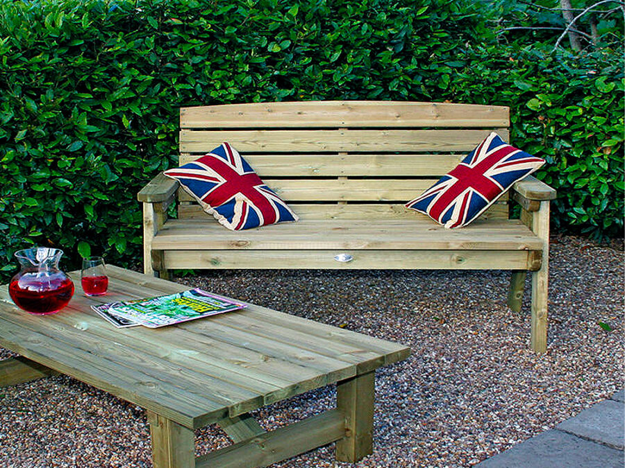 A Hutton Dean 2 Seater wooden garden furniture, with union flag pillows and a display wooden table infront of the seat on a gravel section in a garden setting